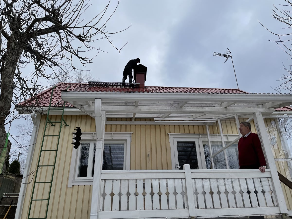 Nuohooja putsaa katolla hormia, toinen mies seisoo mökin terassilla.