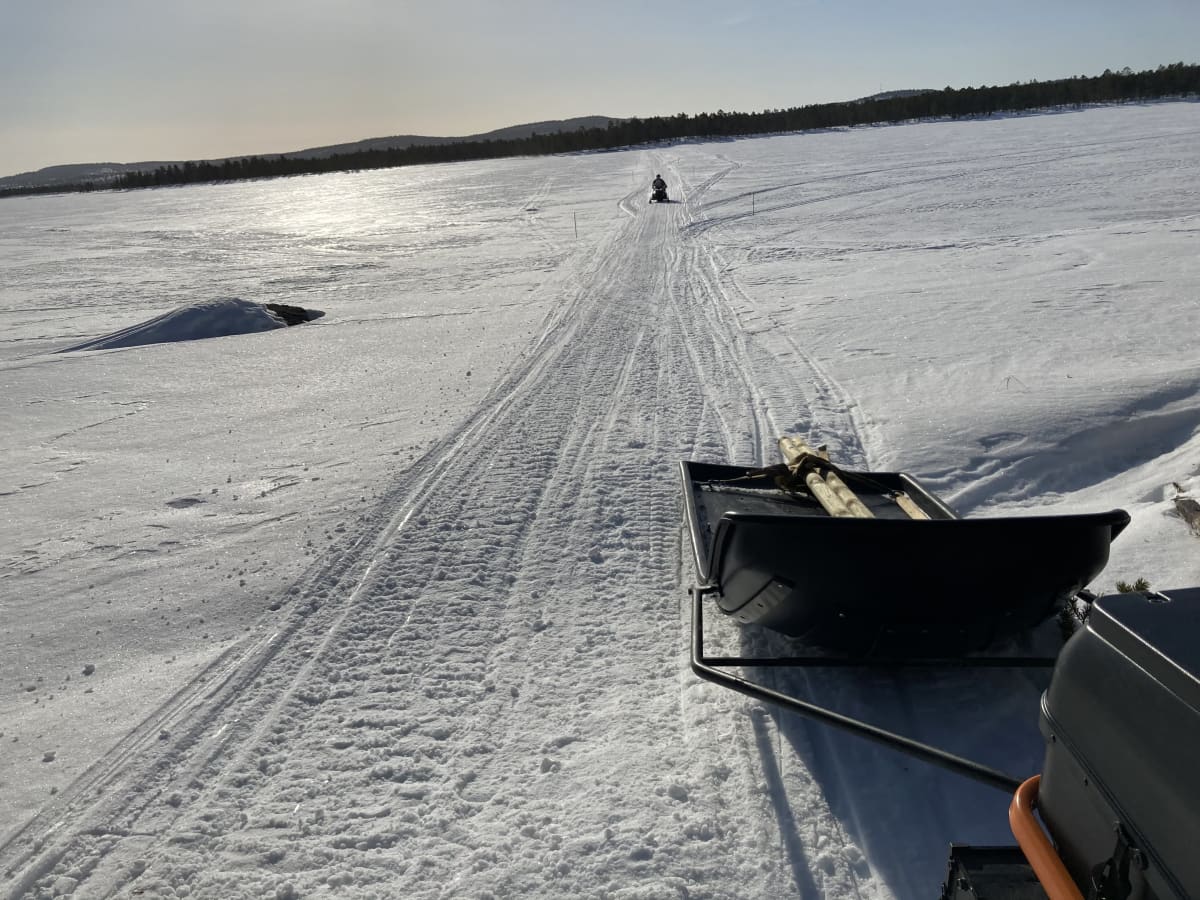 Moottorikelkka ajaa lumisella Inarijärven jäällä. Aurinko paistaa.  