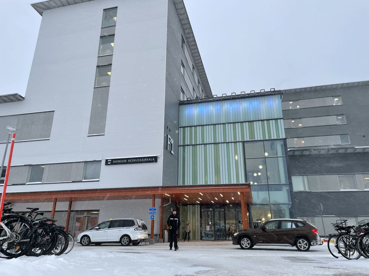 Kainuun keskussairaalan pääsisäänkäynti lumisella kelillä. Mustiin pukeutunut mies kävelee tien yli poispäin sairaalasta.