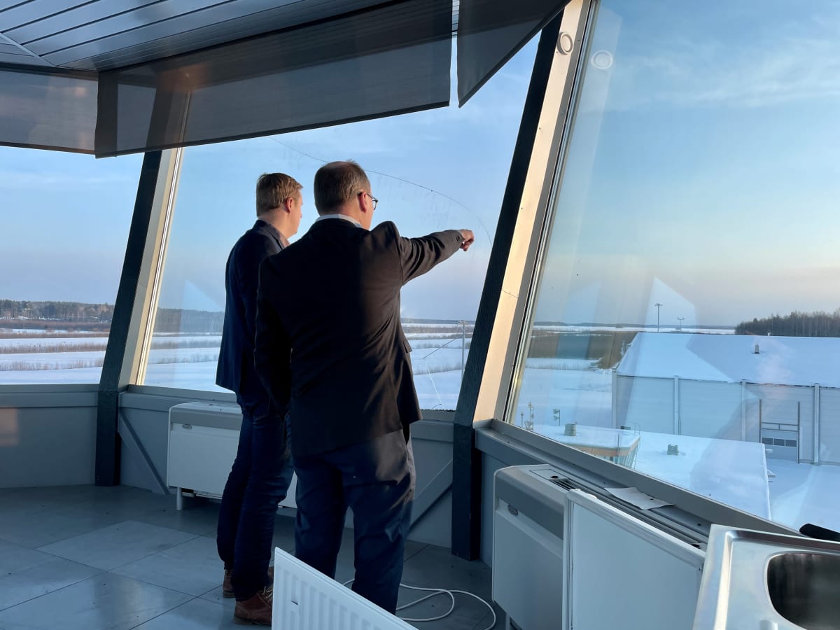 Kaksi miestä seisoo korkeassa lennonjohtotornissa ja katsoo aurinkoista maisemaa.