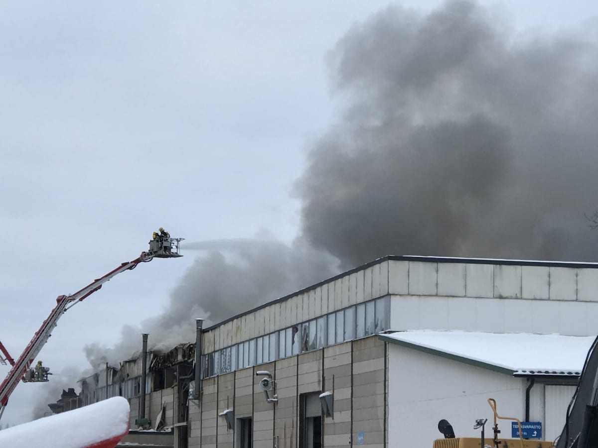 Teollisuushallin katolta nousee tummaa savua. Nosturin korista ruiskutetaan vettä katolle. 