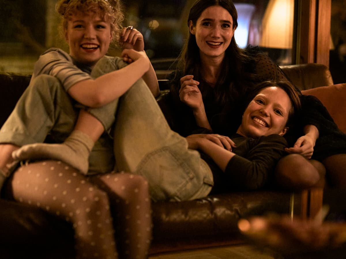 Kuvassa kolme tyttöä istuu sohvalla sylikkäin ja nauraa.