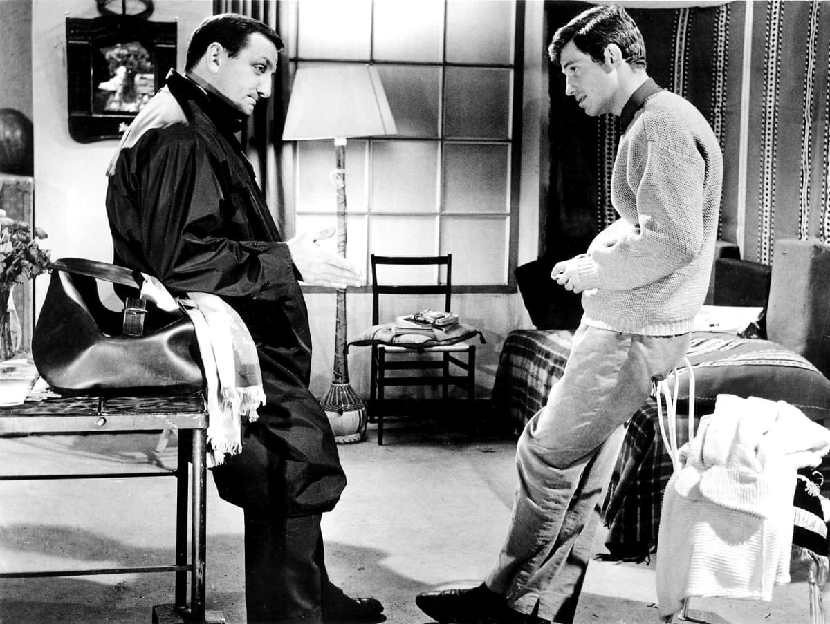 Ventura ja Belmondo seisovat huoneessa vastakkain ja katsovat toisiaan. Ventura nojaa pöytään ja Belmondo tuoliin. Pöydällä Venturan vieressä on nahkainen laukku. Venturalla on tummat vaatteet, Belmondolla vaalea villapaita ja vaaleat housut.