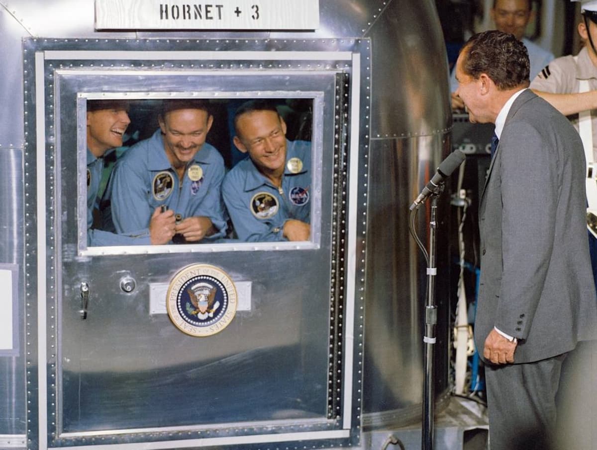Arnstrong, Collins ja Aldrin kurkistevat hymyillen karanteenihuoneen ikkunasta, jonka edessä seisoo Nixon. 
