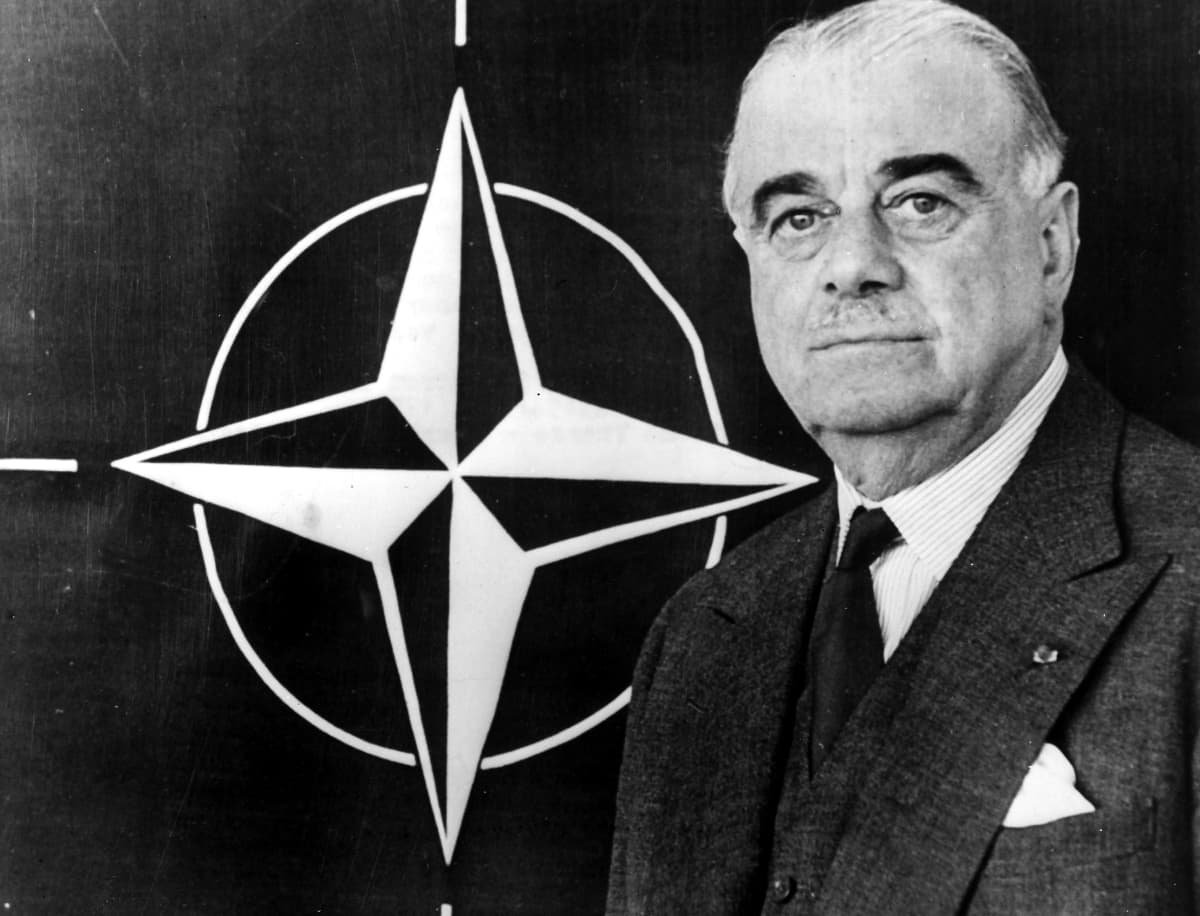 Mustavalkokuvassa Naton logo ja sen vieressä H.L. Ismay, jolla on harmaa puku ja pienet vaaleat viikset. 