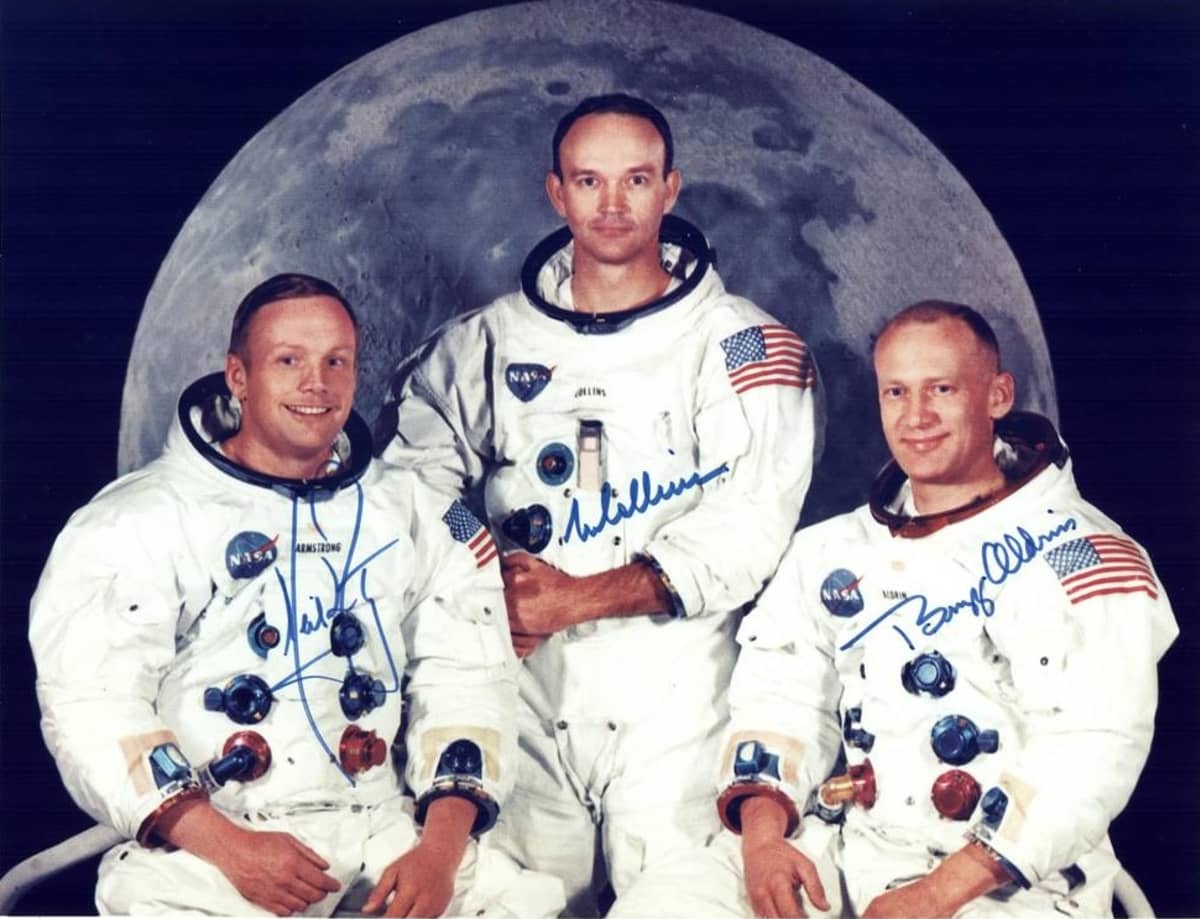 Kolme avaruuspukuista astronauttia Kuuta esittävän kuvan edessä.