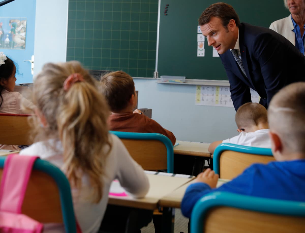 Presidentti Emmanuel Macron kumartuneena puhumaan pulpeteissa istuville pienille koululaisille.