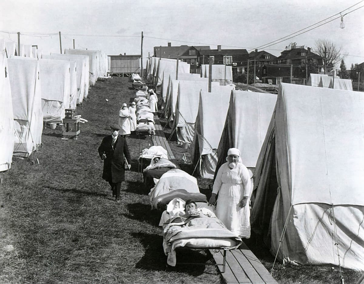 Kentällä on rivi telttoja ja telttojen edessä kenttävuoteilla makaa sairaita. Heidän vieressään seisoo sairaanhoitajia. Tummapukuinen mies kulkee telttojen välissä kulkevalla polulla.