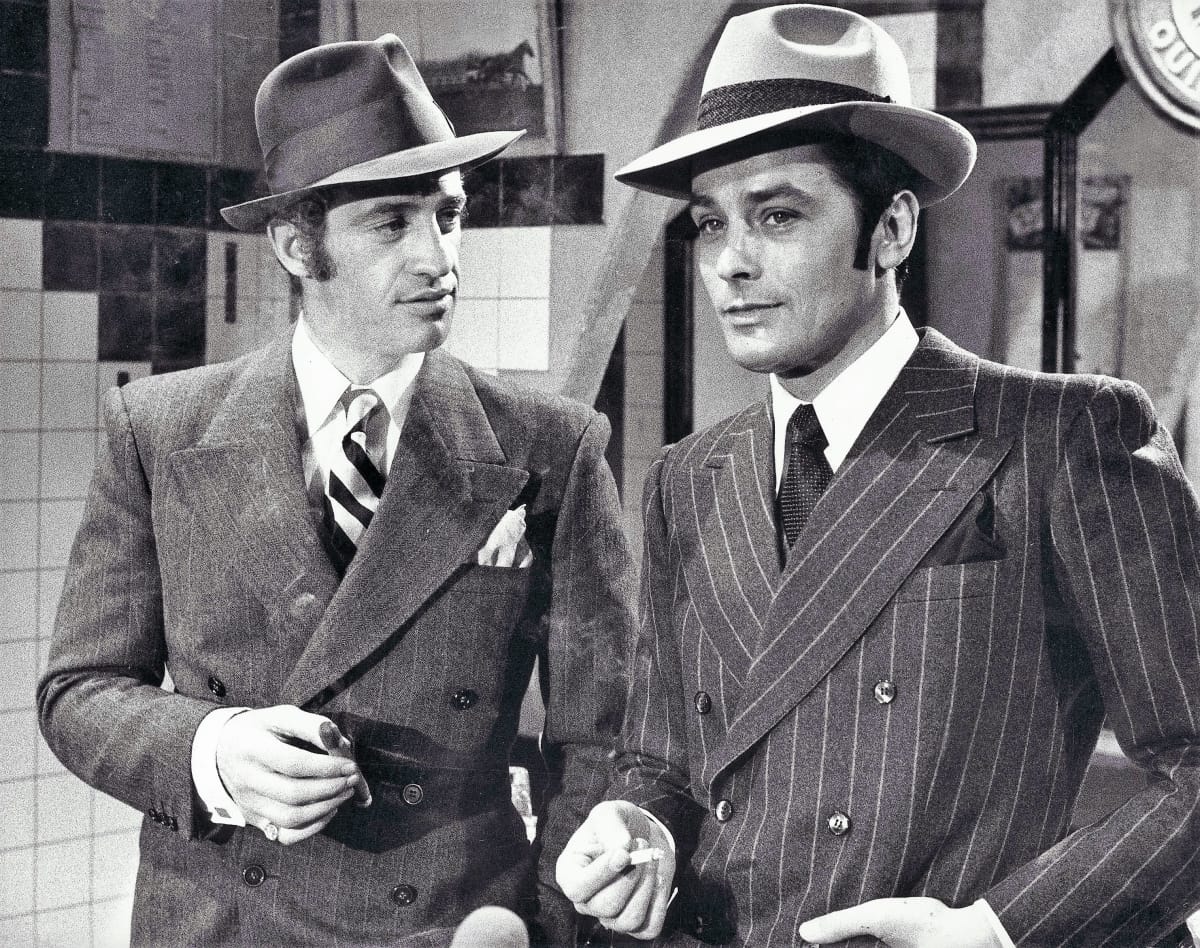 Mustavalkokuvassa Belmondo ja Delon seisovat liituraitaisissa puvuissa tupakat käsissään, huopahatut päässään baarissa. Belmondo katsoo Delonia ja Delon salia kohti. Miehet hymyilevät vienosti.