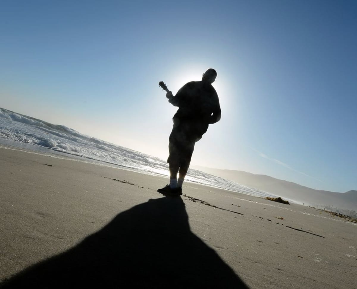 Mies soittaa kitaraa rannalla aurinkoa kohti.