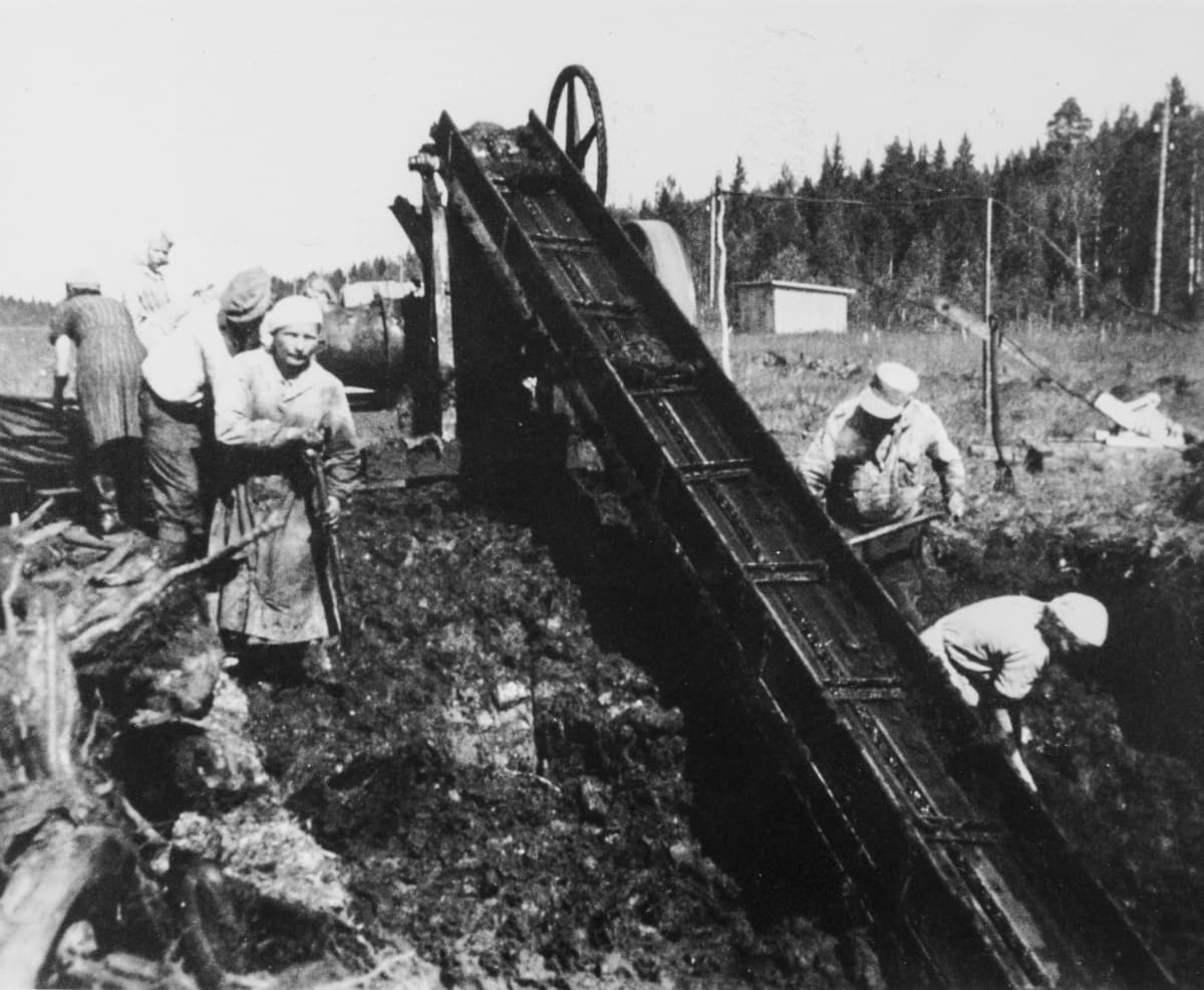 Vanhassa mustavalkokuvassa ihmiset työskentelevät jonkinlaisen koneen kanssa ulkona. 