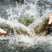 Poika hyppäsi veteen selällään vain jalkapohjat näkyvät pärskeiden seasta.
