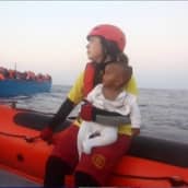 Nainen lapsi sylissään veneessä, taustalla ihmisiä täynnä oleva vene. 