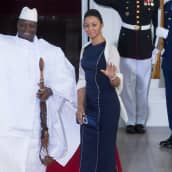 Jammeh ja hänen puolisonsa seisovat punaisella matolla ja tervehtivät ihmisiä hymyillen.