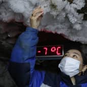 Maskiin ja talvitakkiin pukeutunut tutkija koskettelee katon jäämuodostumaa. Taustalla lämpömittari näyttää seitsemää pakkasastetta. 