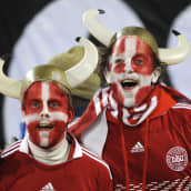 Kaksi huutaavaa nuorta miestä, joilla on Tanskan joukkueen punainen takki, kasvot maalattu Tanskan lipun värein ja päässä movinen sarvikypärä. 