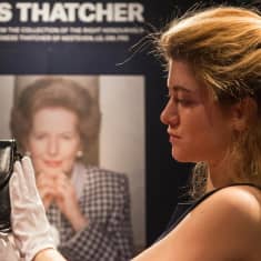 Huutokauppakamarin työntekijä pitelee hansikoiduin käsin pientä mustaa käsilaukkua. Taustalla juliste, jossa Thatcherin kuva ja nimi. 