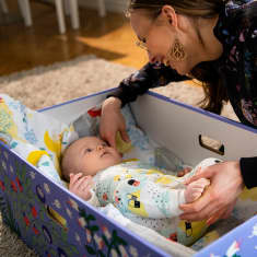Nainen hymyilee vauvalle, joka makaa äitiyspakkauslaatikossa.