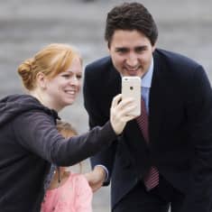 Justin Trudeau ja naiskannattaja katselevat hymyilleen valokuvaa naisen kännykästä. Naista vasten painautuu pieni tyttö. 