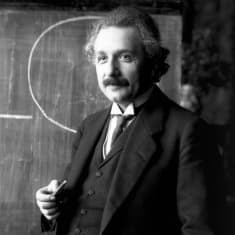 Liivipukuun pukeutunut Einstein liitutaulu  edessä. 