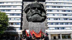 Pieni joukko nuoria ihmisiä punaisten banderollien kanssa makailee ja istuskelee jyhkeän betonisen Marxin päätä esittävän patsaan jalustan juurella. Taustalla vaalea DDR-tyylinen kerrostalo.
