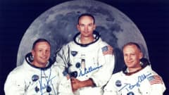 Kolme avaruuspukuista astronauttia Kuuta esittävän kuvan edessä.