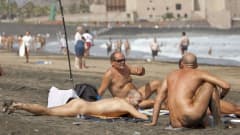 Kolme alastonta miestä lekottelee rannalla. 