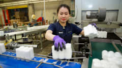Työntekijä siirtelee käsin kuuden pöytäkynttilän pakkauksia hihnalta toiselle.