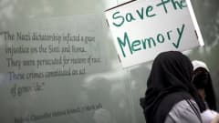 Mielenosoittaja pitelee pahvilaatikosta tehtyä kylttiä, jossa lukee englanniksi "pelastakaa muisto". Taustalla näkyy muistomerkin teksti, jossa liittokansleri Helmut Kohl tunnustaa romanien kansanmurhan. 
