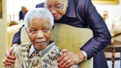 Mandela istuu tuolissa, Machel kumartuneena pitelee hänen olkapäistään kiinni.