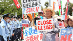 Rivi sotilaita kasvosuojuksissaan tarkkailee iäkkäitä mielenosoittajia, joiden kylteissä vaaditaan englanniksi ja japaniksi, että kaikki tukikohdat suljetaan. 