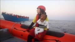 Nainen lapsi sylissään veneessä, taustalla ihmisiä täynnä oleva vene. 