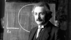 Liivipukuun pukeutunut Einstein liitutaulu  edessä. 