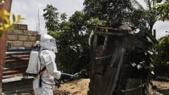 Suojavarusteisiin pukeutunut työntekijä ruiskuttaa desinfiointiainetta ebola-potilaan puutarhavajaan.