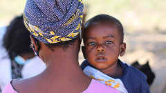 Selin oleva nainen kantaa sylissään pientä lasta, joka katsoo hänen olkansa ylitse vakavana kuvaajaa silmiin. 