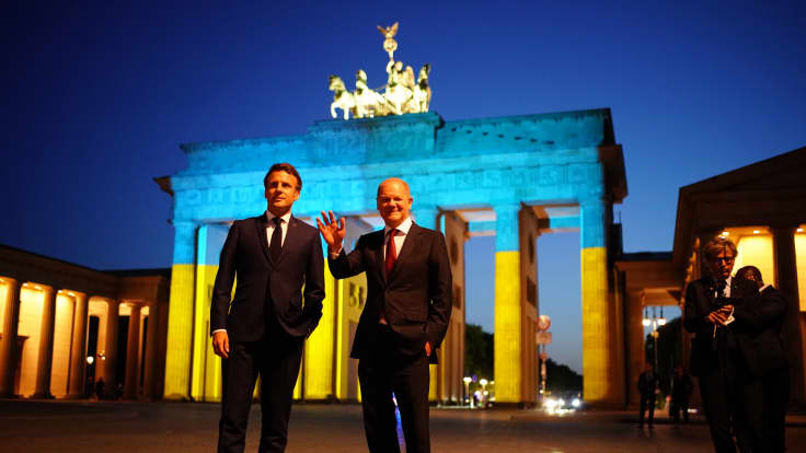 Kaksi tummiin pukeutunutta miestä ison keltasinisen portin edessä pimeässä illassa. Olaf Scholz oikealla vilkuttaa kameroille.