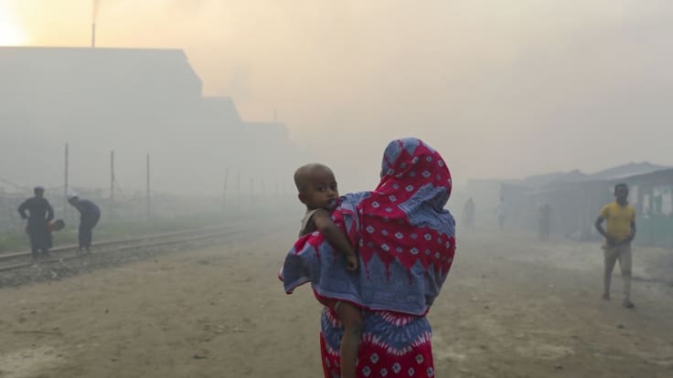 Nainen kantaa pientä lasta ilmansaasteiden sumentamassa maisemassa rautatien varressa.