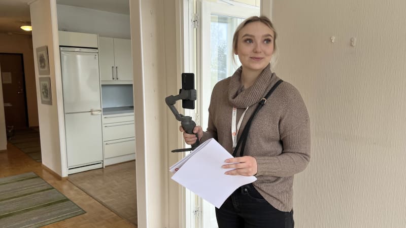 Менеджер з продажу OP Koti Päijät-Häme Ііда Саастамойнен віртуально представляє житловий будинок клієнтам за допомогою мобільного телефону.  У Іїди в руці підвіс, до якого прикріплений мобільний телефон, а в іншій руці папери.