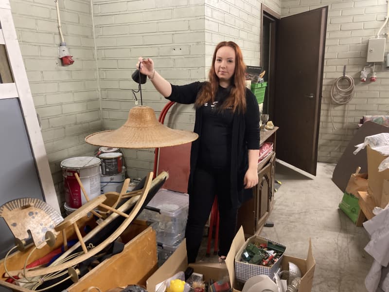 A woman holds a rattan ceiling lamp in a storage room full of stuff.