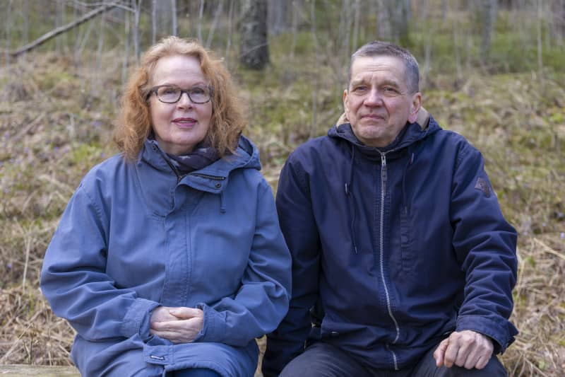 Riitta (left) and Olli Jalonen sit on a bench in the forest and look at the camera.