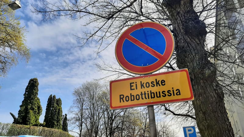 A round traffic sign with a red crossed line on a blue background and a yellow-red sign below that reads Not for Robot Bus.  In the background, treetops and a blue sky with thin clouds.