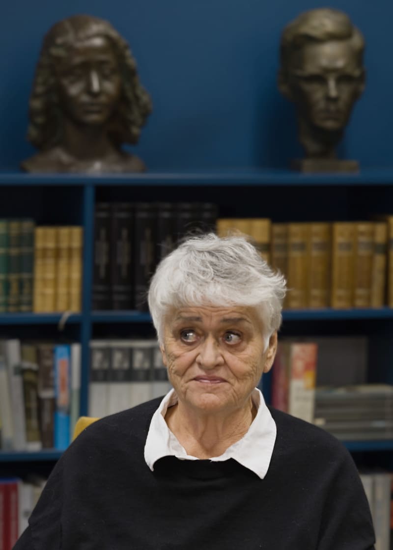 Піркко Саісіо сидить перед книжковою полицею й дивиться на сторінку.  На задньому плані на книжковій полиці видно дві письменницькі ручки.