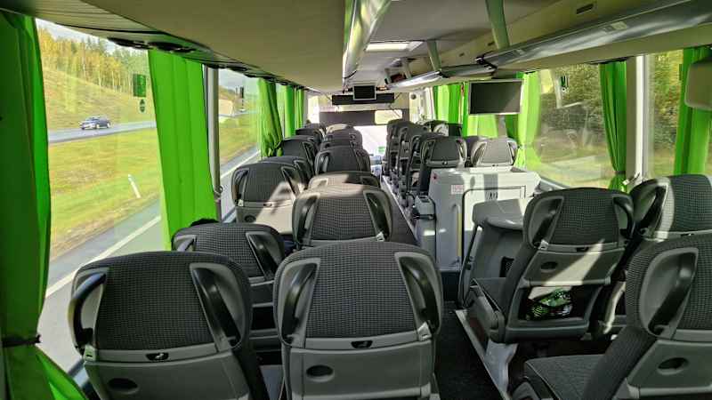 The picture shows the interior of a FlixBus bus.  The coach has seats with individual backrests, green curtains on the windows and some passenger amenities such as a toilet and a mobile phone charging socket.  The picture was taken from the back of the bus towards the front, and shows the aisle and several rows of two seats.