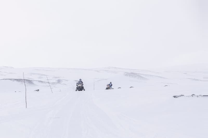 Two snowmobilers driving on a snowy desert, with a snowy sky covering the landscape.