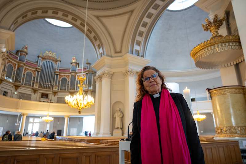 An older woman in the priest's outfit stands in church.  In the background you can see decorative arcs and spectacular organ on the loft.
