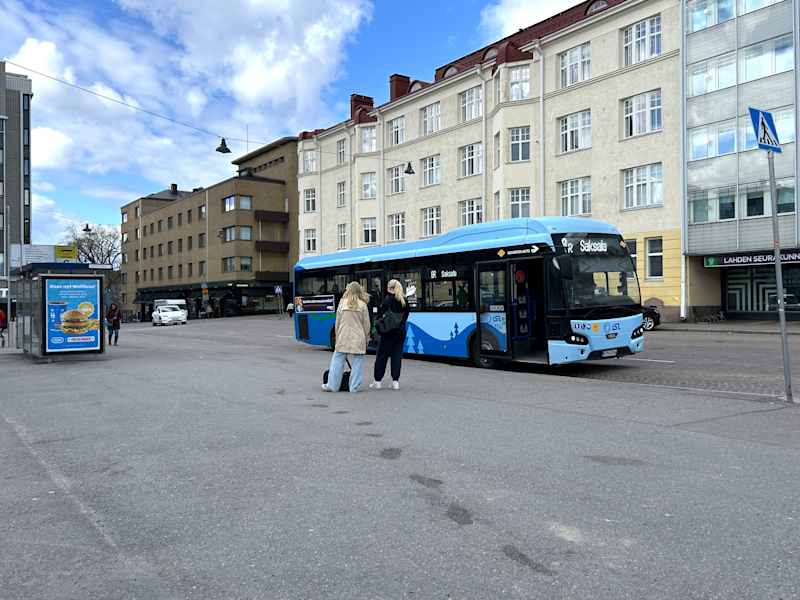Two people stand next to the bus stop.  There is a blue bus parked on the street.