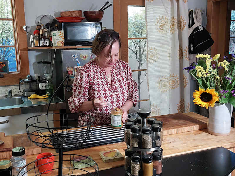 A woman takes a peanut butter from a jar in a bright kitchen with a flower arrangement.