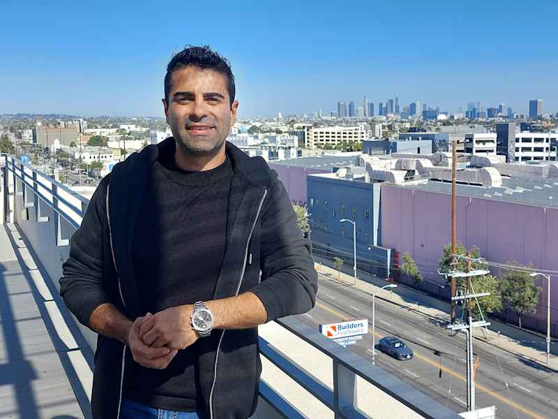 A man with a black shirt and a hoodie looks at the camera on a narrative roof terrace.  In the background is Los Angeles Downtown.