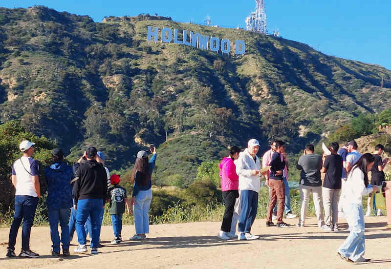 Tourists stand in front of a steep slope and has a Hollywood sign at the top.
