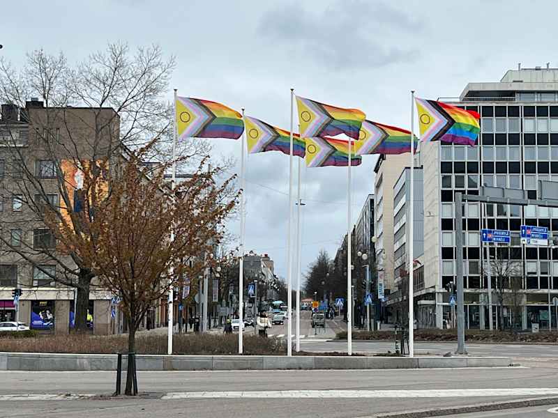 The picture shows colorful Pride tickets fluttering in the flag rods in the urban landscape.  There are gray concrete buildings and streets in the background.